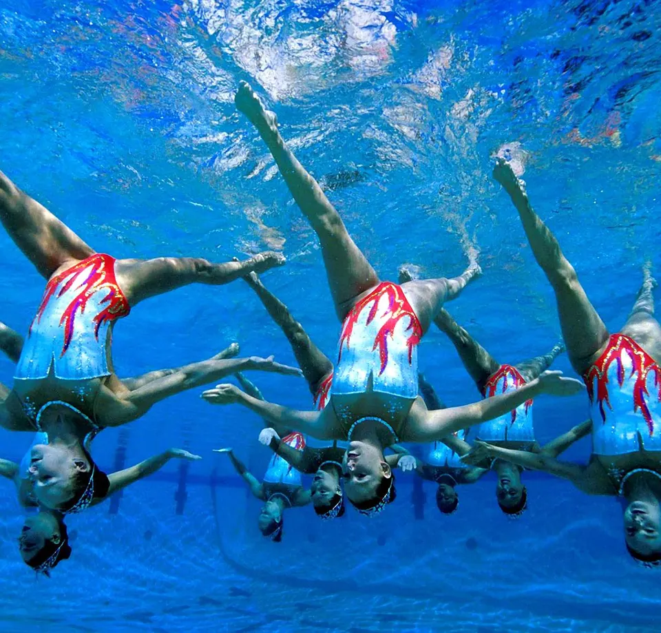 An artistic swimming team upside down underwater in matching red and white costumes, legs raised in unison
