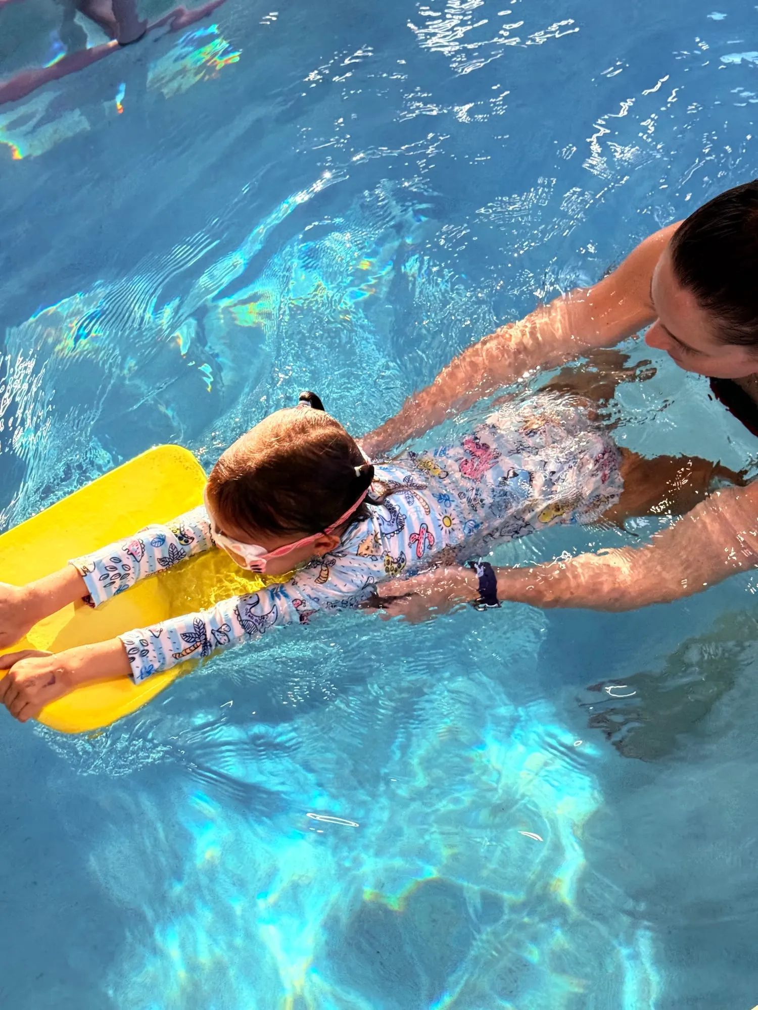 Three children in swim caps and goggles grinning up from the pool wall, kickboards in their hands