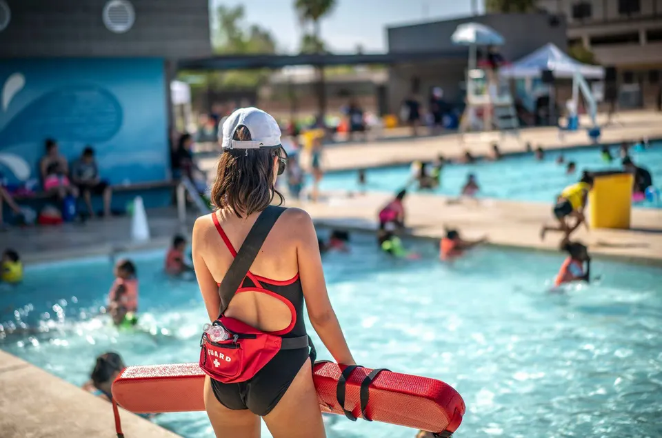 A lifeguard in a red and black suit standing at the edge of a busy outdoor pool, rescue tube under one arm and a hip pack marked GUARD
