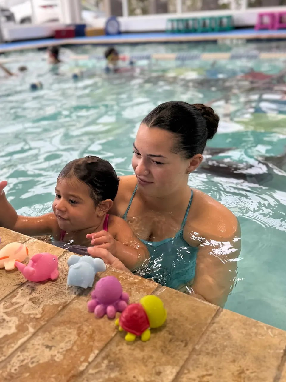 An adult in the water holding a toddler at the pool wall, bath toys lined up on the edge