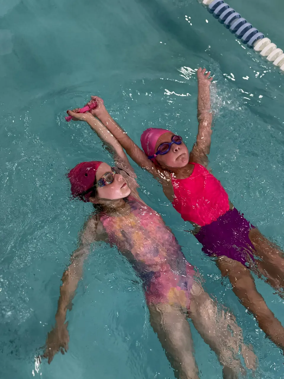 Two girls swimming backstroke side by side down the lane, faces up and relaxed
